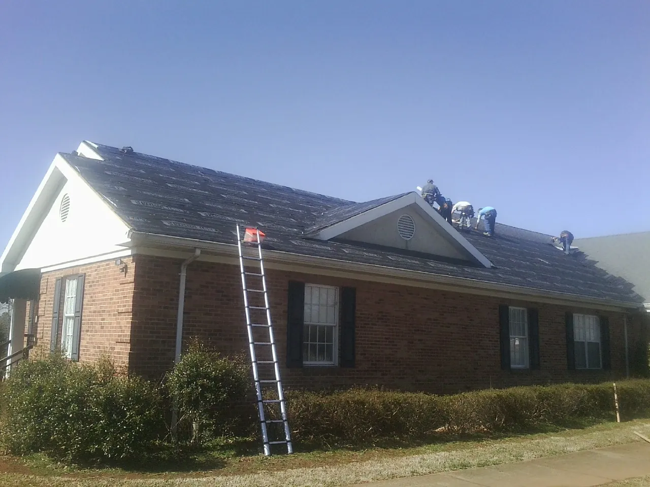 Crew tearing shingles off a brick ranch roof with a ladder set — Emergency Roof Repair work by our crew in Hatton