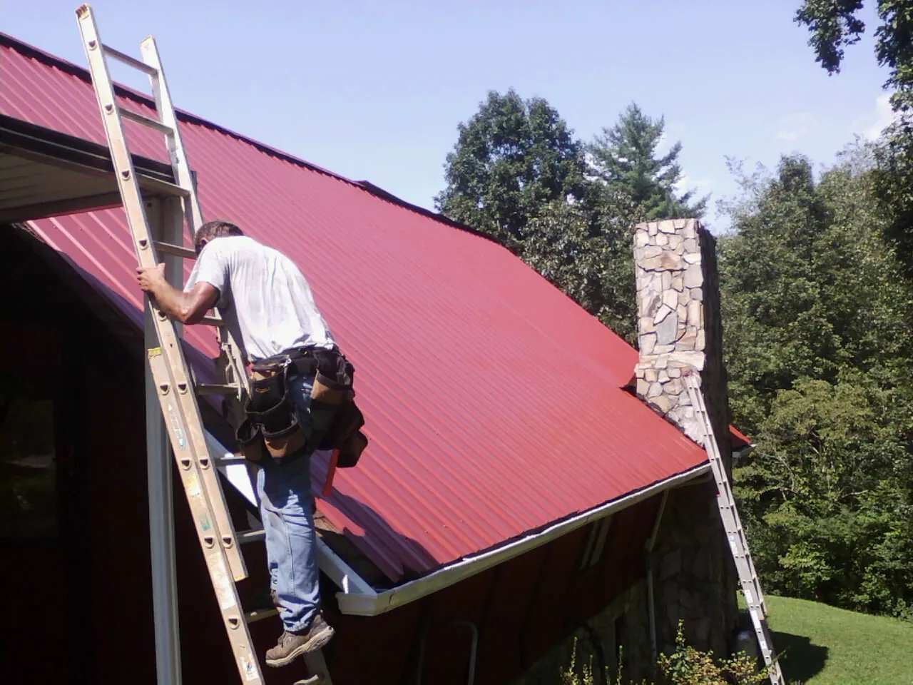 Roofer on a ladder beside a red metal roof and stone chimney — Soffit & Fascia Repair by Roofing Brothers in Hatton
